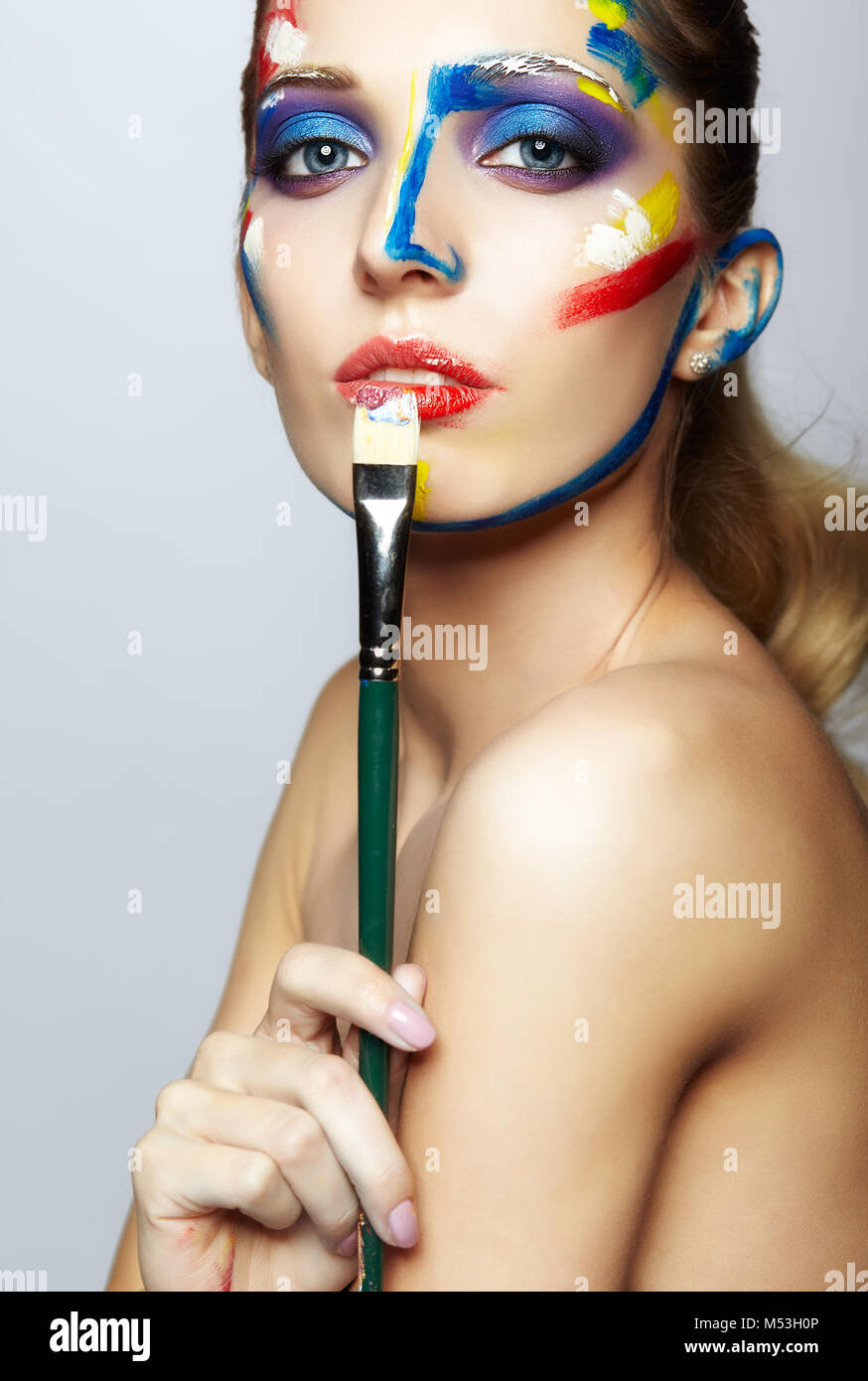 Young woman painter with paint brush in hand and acrylic paint on face ...