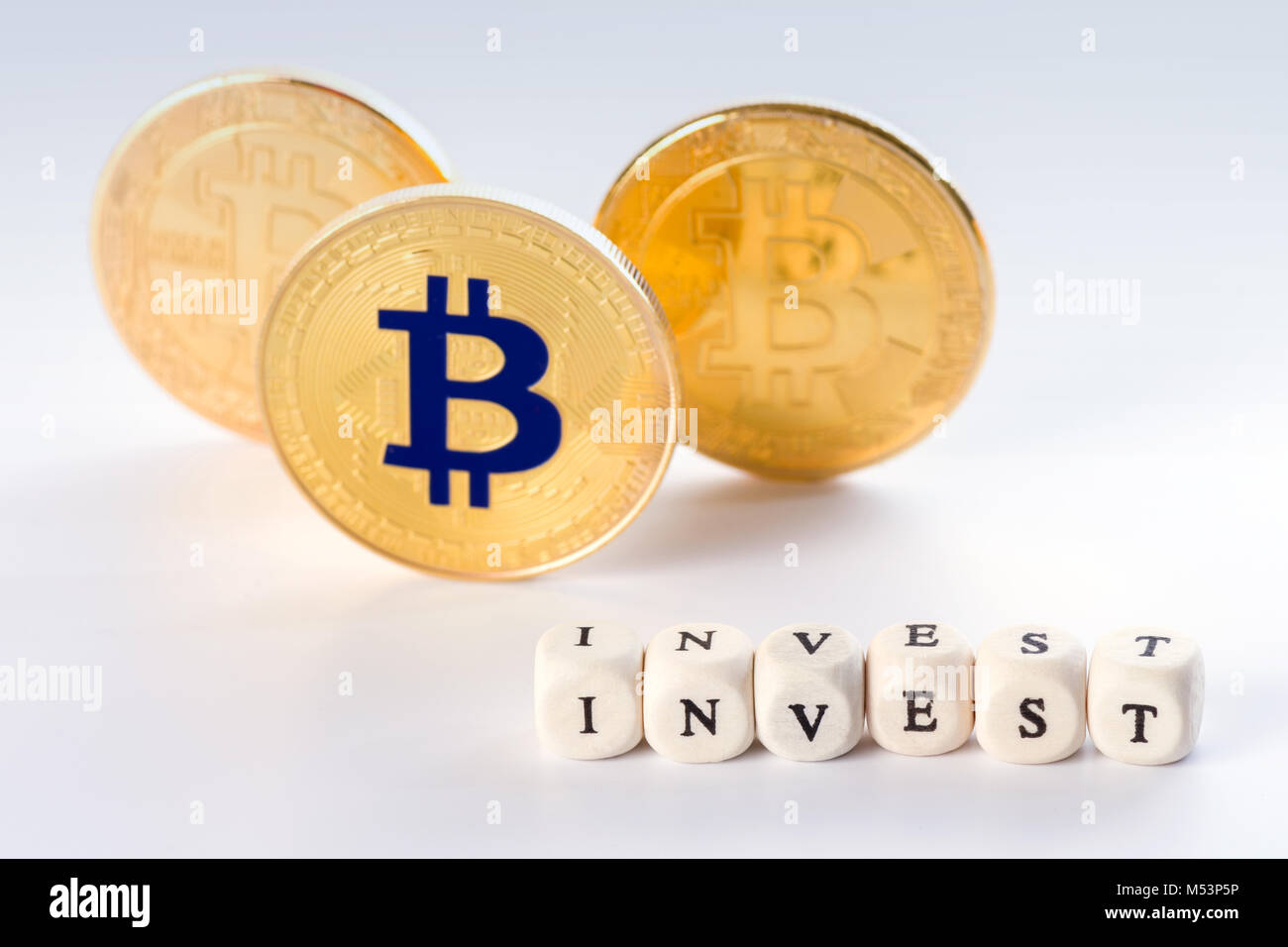 The word INVEST in sharp focus made of wooden blocks and three standing bitcoin coins behind it. Investing in crypto currency concept on white backgro Stock Photo
