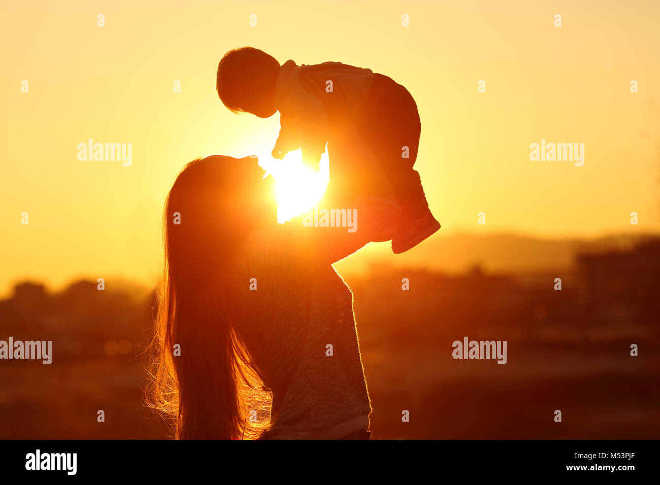 Backlight silhouette of a mother raising her kid son at sunset with the sun in the middle Stock Photo
