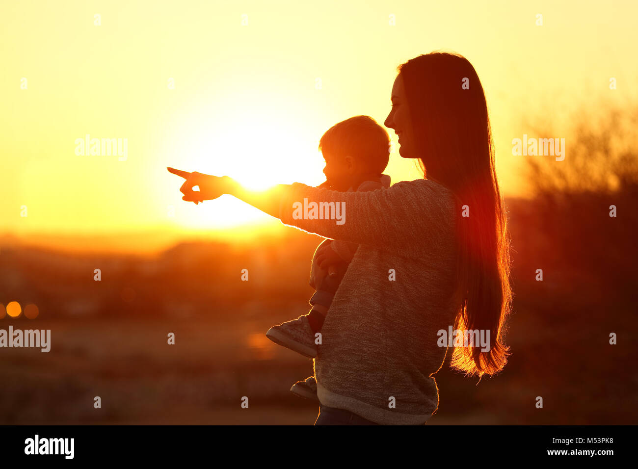Backlight silhouette of a mother pointing horizon with her baby at sunset Stock Photo