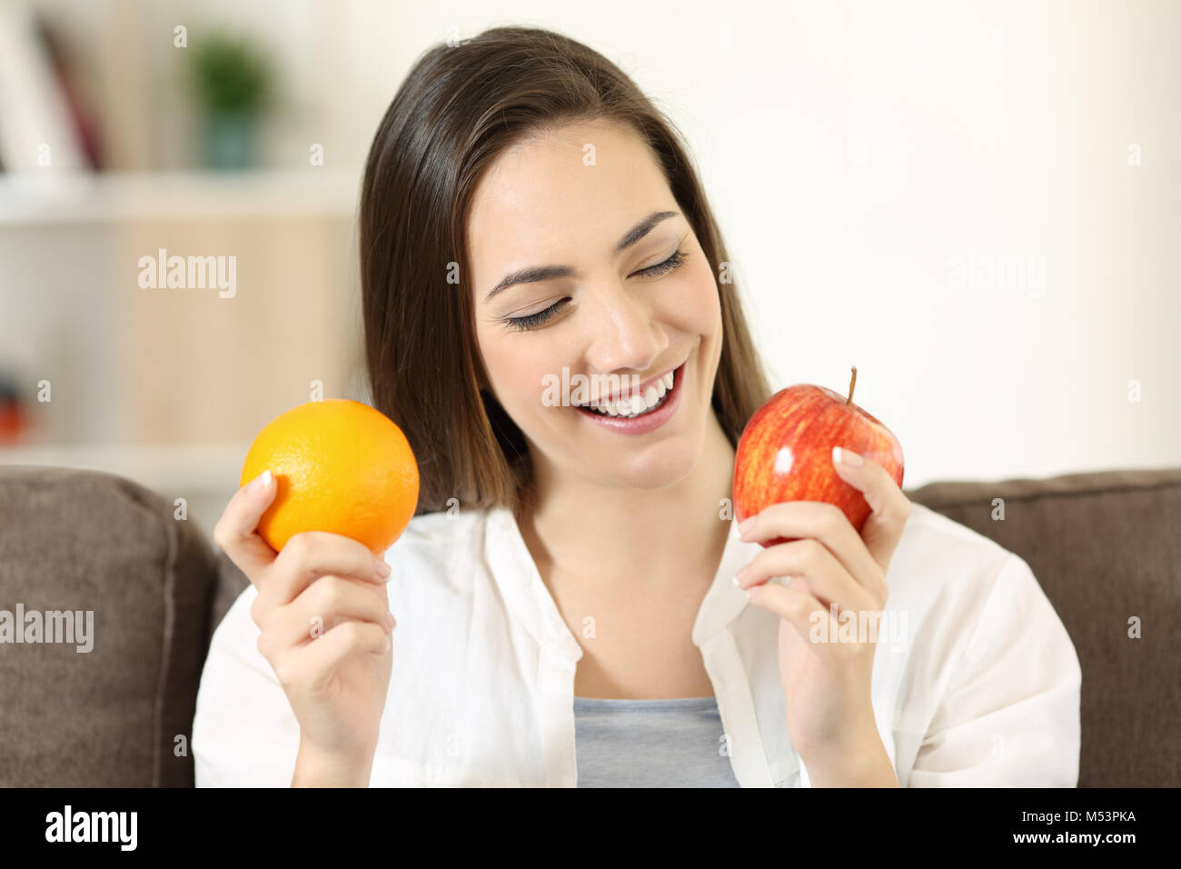 Front view portrait of a happy woman deciding between orange and apple sitting on a couch in the living room at home Stock Photo