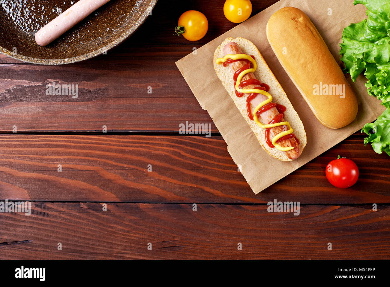 Hot Dog on craft paper bag with sausage in pan, bun, tomatoes and salad flat lay on wooden background. Top view. Stock Photo