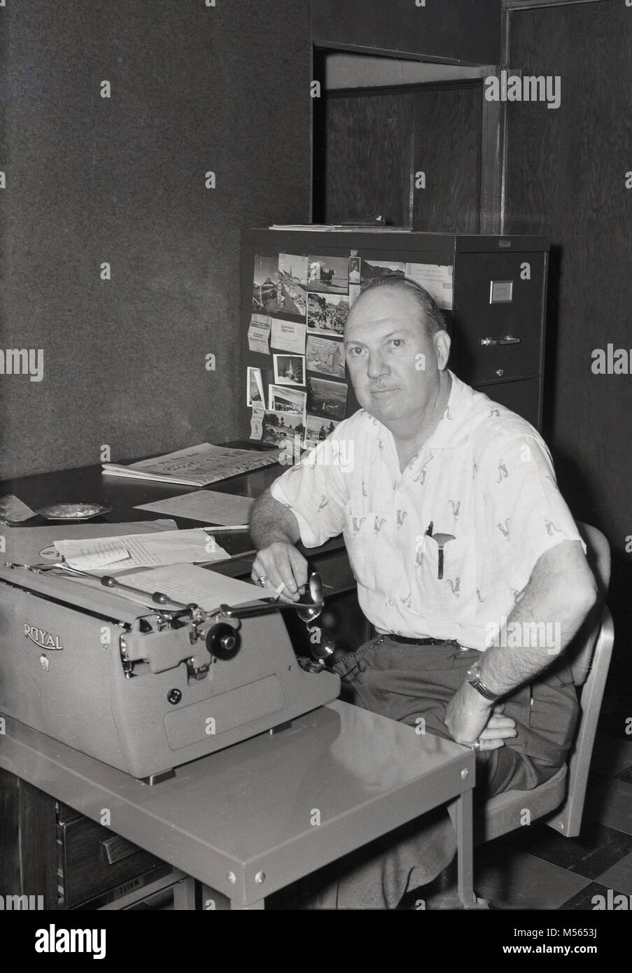 1960s, USA, a middle-aged male journalist in a small office typing up his column using an american made 'Royal' typewriter. The 1960s was a boom time for typewriter sales and usage and the 'Royal' one of its most famous brands, with their first typewriter introduced in 1906. Stock Photo