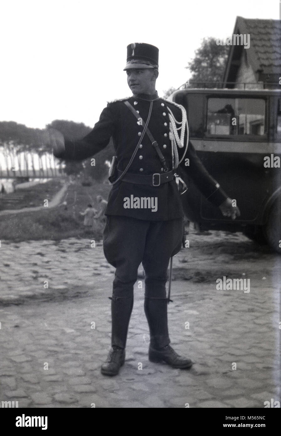 1920s, historical picture showing a young French policeman or 'gendarme' in his decorative military uniform, with tunic and breeches, standing on a village cobbled road using hand signals to direct motor vehicles of the era. The 'Gendarmerie Nationale is part of the French armed forces and has been since 1791. Stock Photo