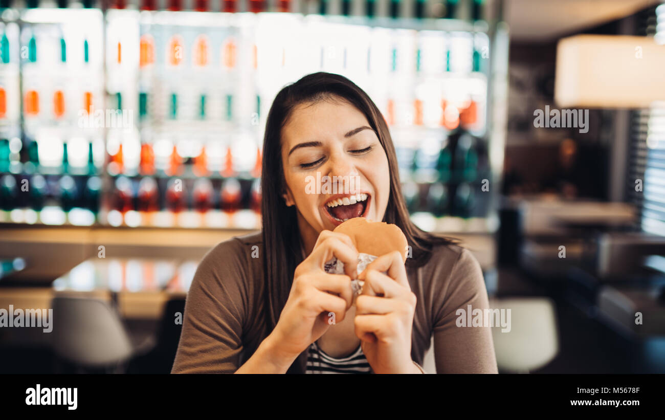 Young woman eating fatty hamburger.Craving fast food.Enjoying guilty pleasure,eating junk food.Satisfied expression.Breaking diet rules,giving up diet Stock Photo