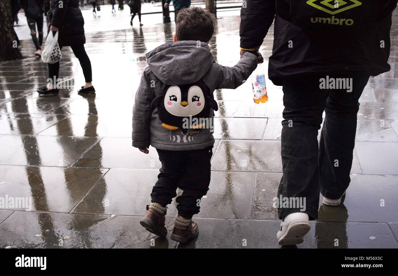 A young boy holds hand with a guardian wearing a penguin back pack walking through the wet streets of London. Stock Photo