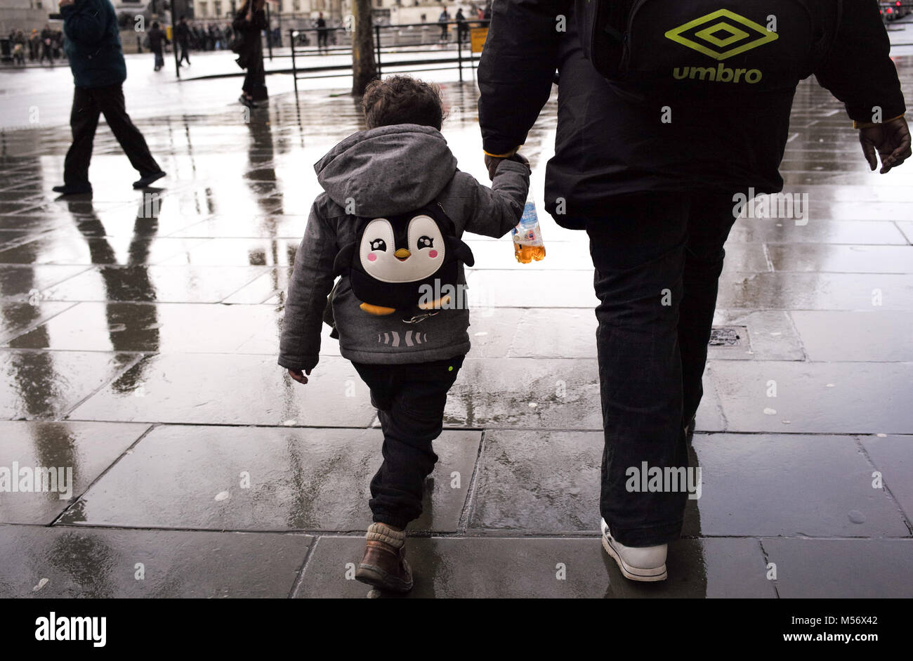 A young boy holds hand with a guardian wearing a penguin back pack walking through the wet streets of London. Stock Photo
