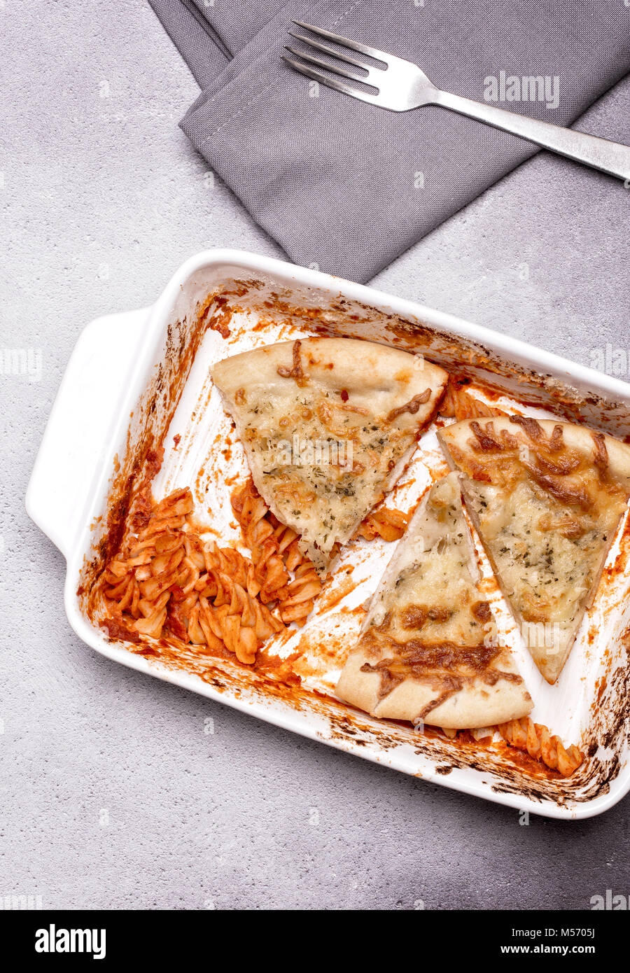 Dish of leftover pasta and cheesy garlic bread with a fork and napkin on a grey rustic background Stock Photo