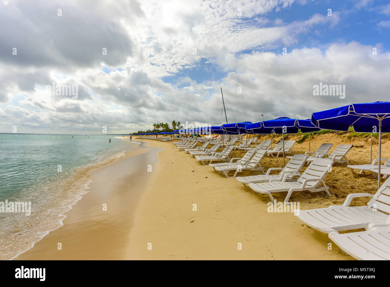 white chaise lounges and blue umbrellas stand on a beach in the sand near the ocean with green waves and a blue sky with white clouds Stock Photo