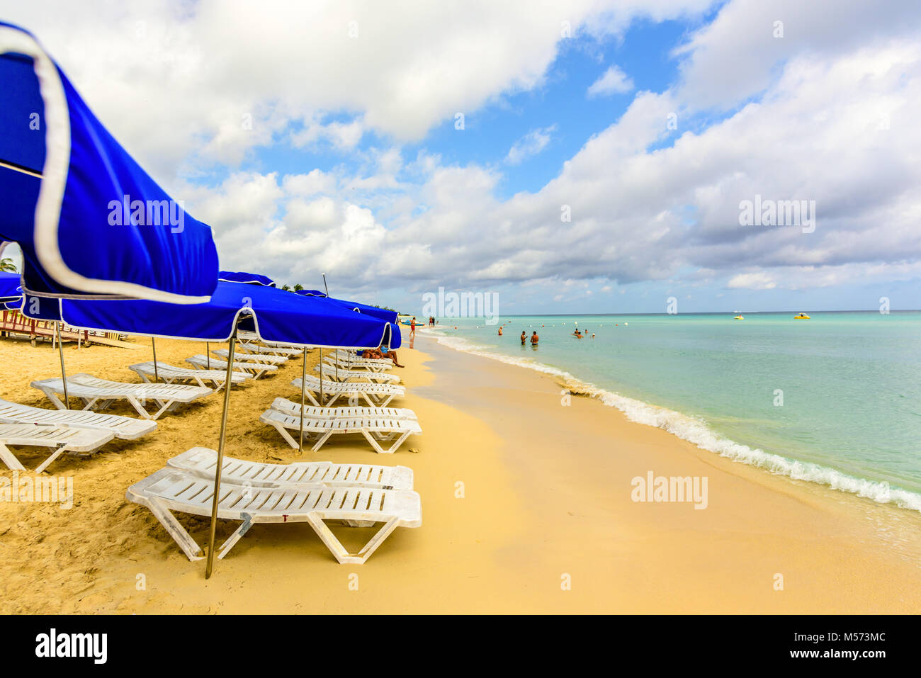 white chaise lounges and blue umbrellas stand on a beach in the sand near the ocean with green waves and a blue sky with white clouds Stock Photo