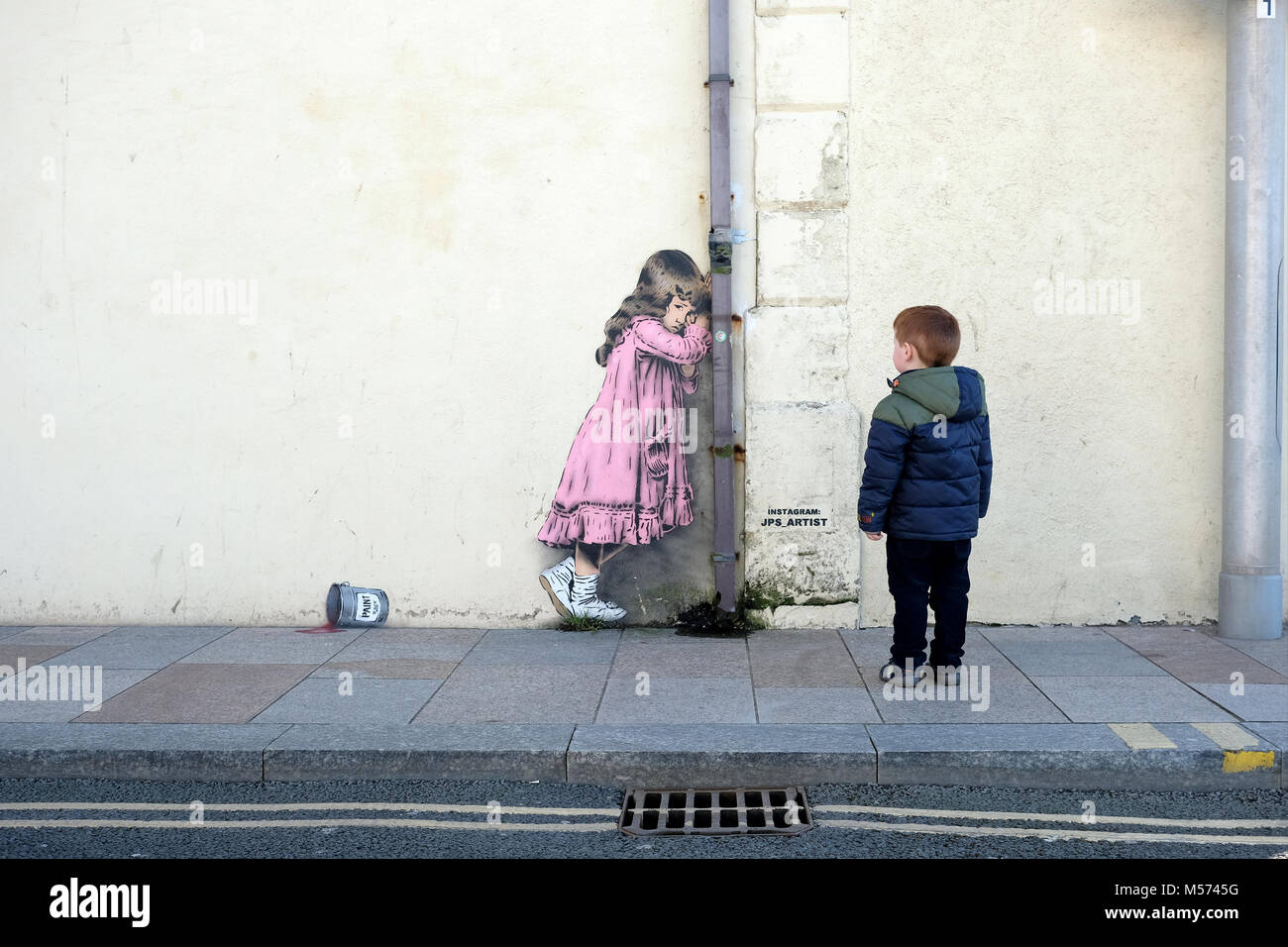 A young boy looks at a banksy style graffiti artwork of a young girl crying over a spilled tin of paint. The artworkby JPS and titled Spilled Anarchy Stock Photo