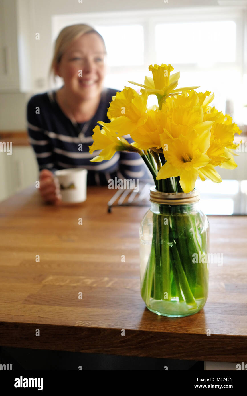 A bunch of freshly picked daffodils arranged in a glass vase placed on a top in a kitchen. A woman is relaxing with a mug of coffee in the background Stock Photo