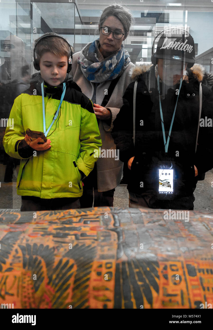 Mother and two sons (listening to audio guide through headphones), looking through glass at a mummy case, at the British Museum, London, England, UK. Stock Photo