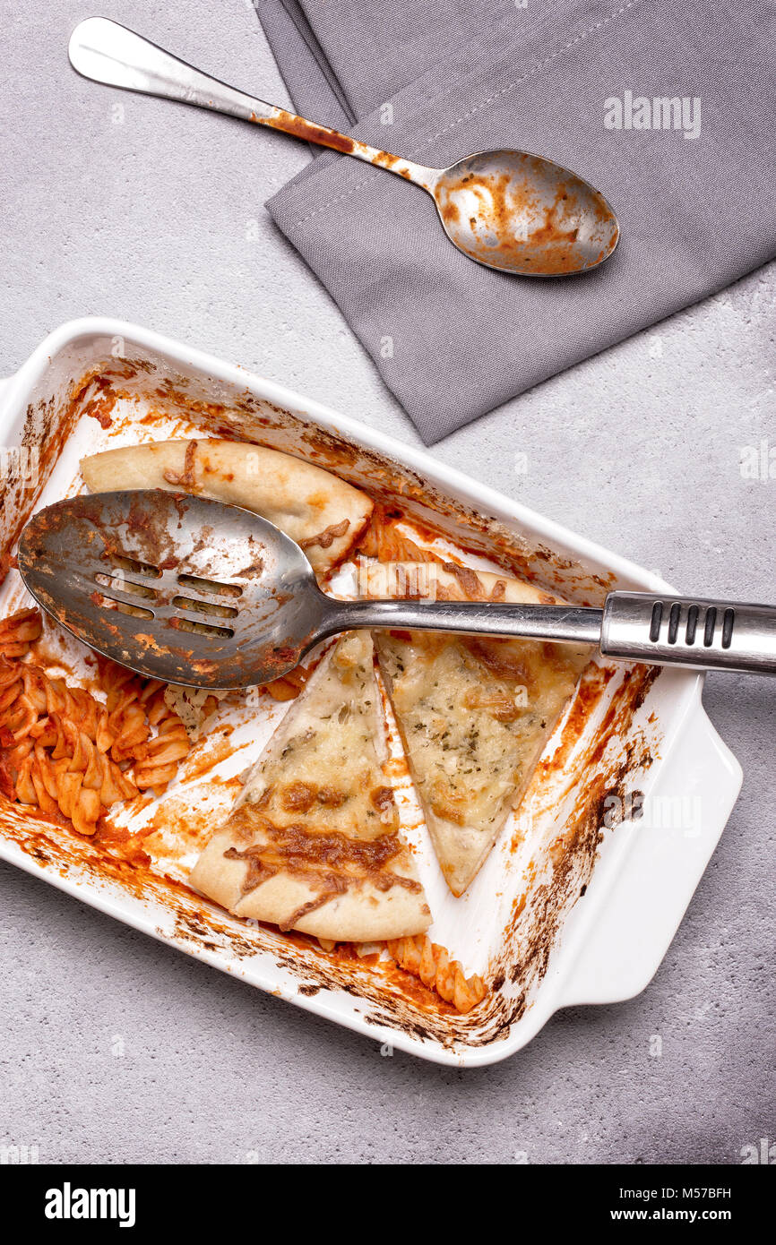Dish of leftover pasta bake and cheesy garlic bread with a spoon and napkin on a grey rustic background Stock Photo