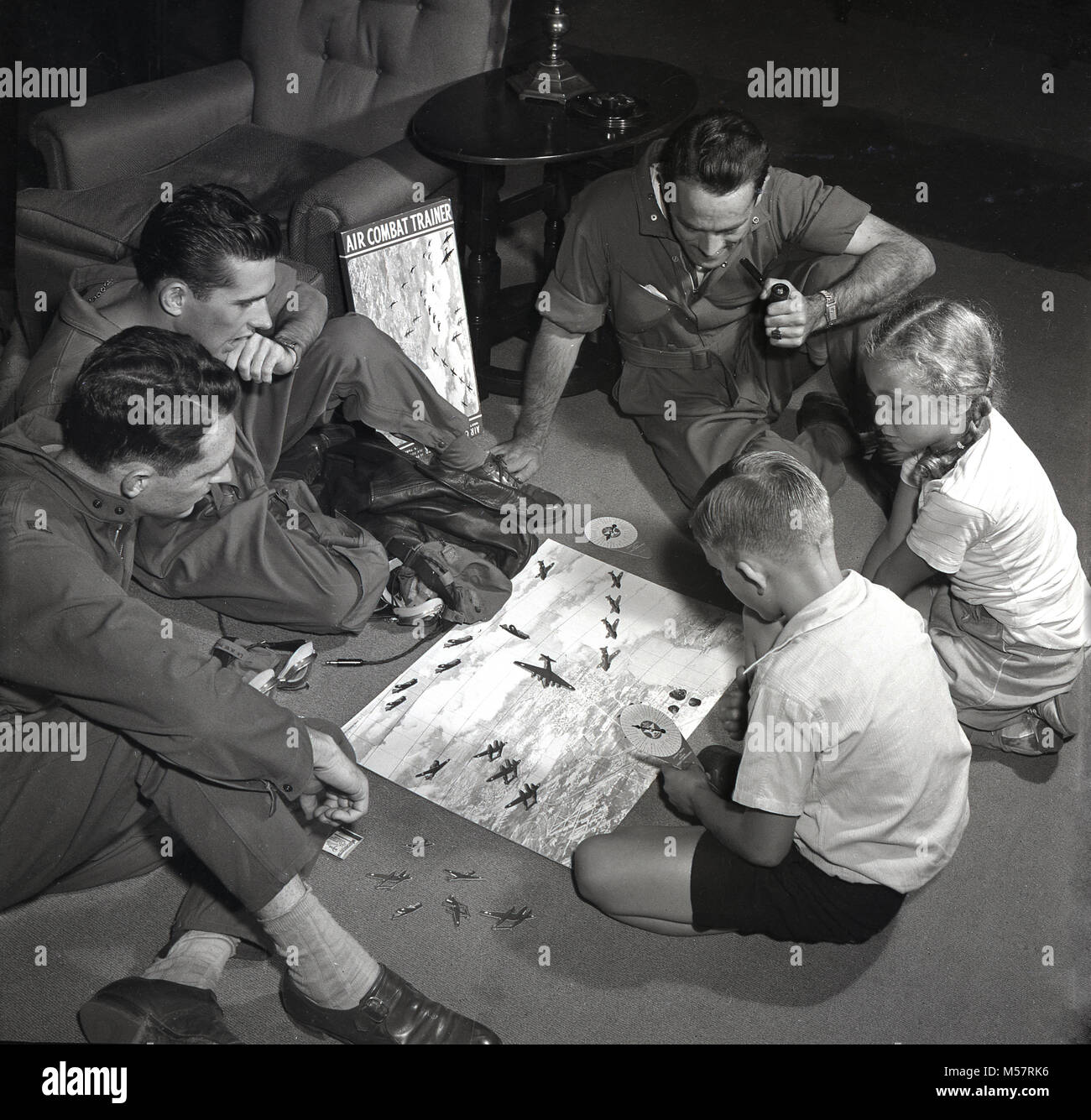 1942, USA, historical, three servicemen of the United States Army Air Forces (USAAF) wearing their flying kit sitting inside playing the new board game, 'Air Combat Trainer' with two young children. Produced in conjunction with the US War Department, the game was a test of skill at combat flying, using miniatures of war planes and combat manoeuvres. Stock Photo