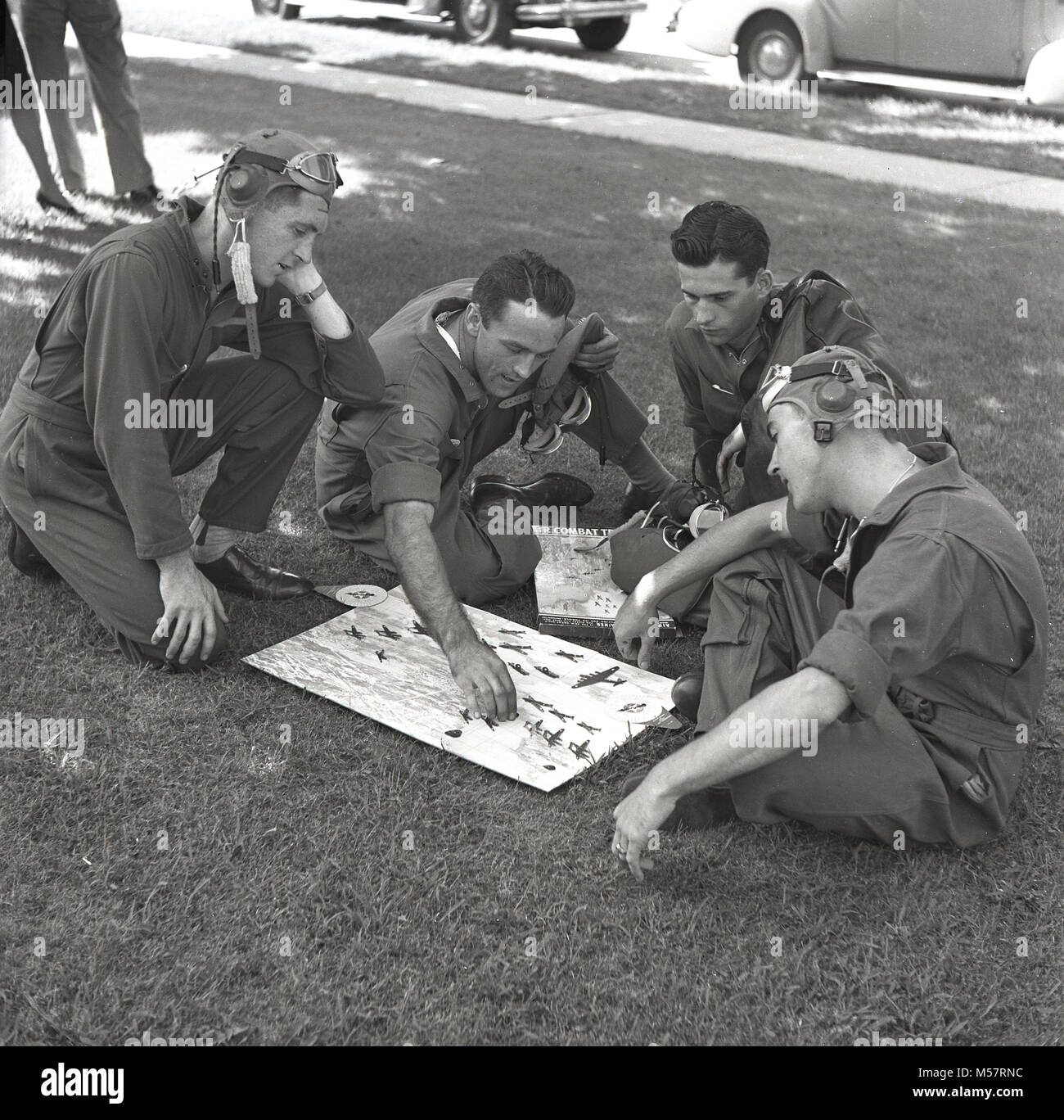 1942, USA, historical, four servicemen of the United States Army Air Forces (USAAF) wearing their flying kit sitting on a grass verge outside playing the new board game, 'Air Combat Trainer'. Produced in conjunction with the US War Department, the game was a test of skill at combat flying, using miniatures of war planes and combat manoeuvres. Stock Photo