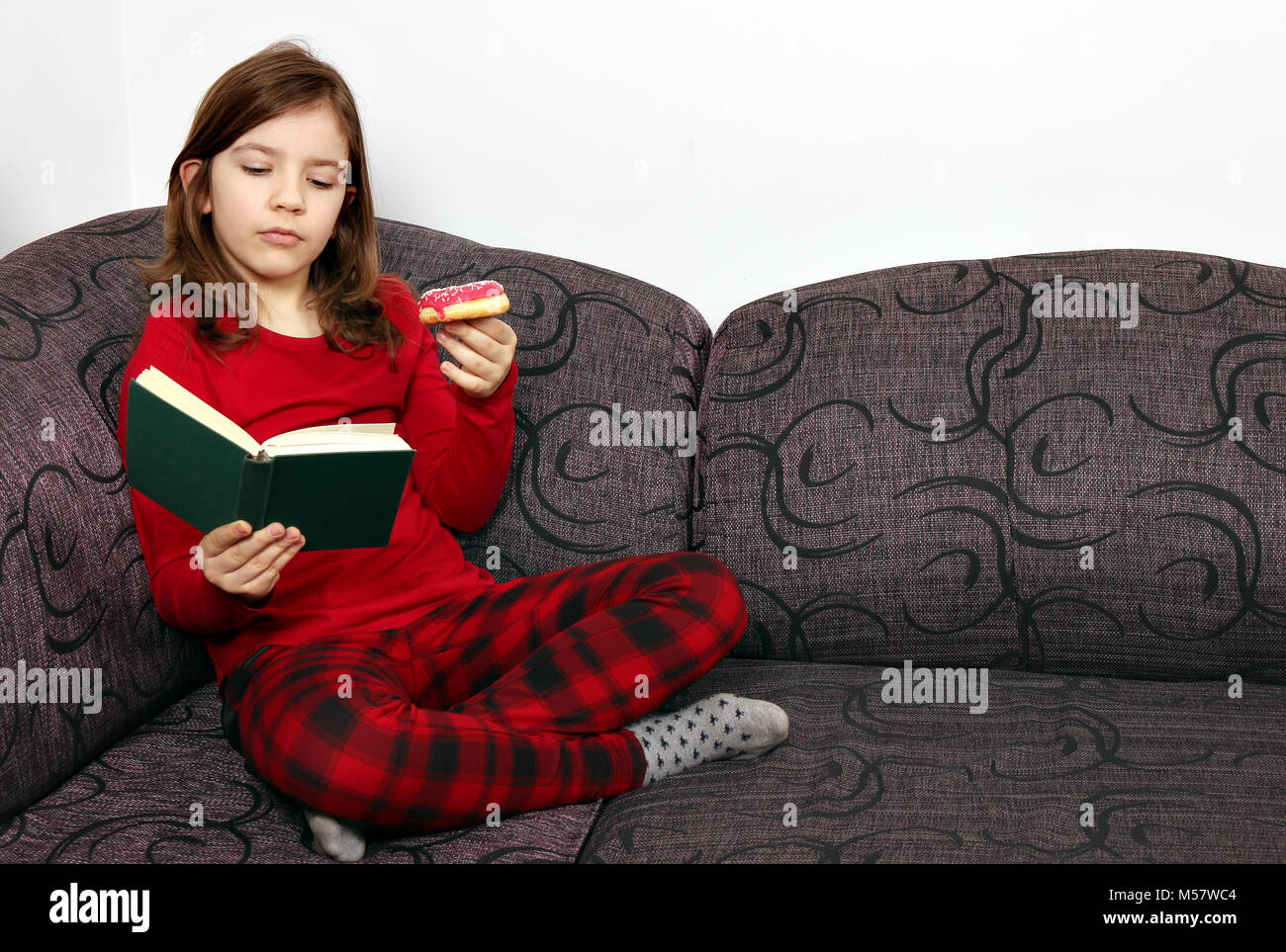 Little girl reading a book and eating a donut at home Stock Photo