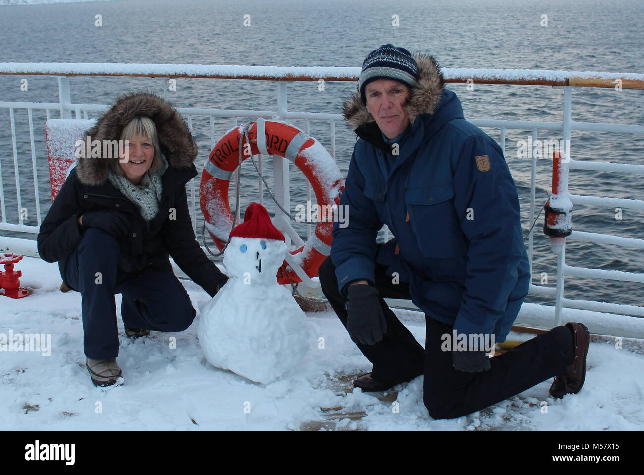 Stowaway snowman joins couple wearing parkas on Hurtigruten winter cruise in the fjords in Norway with lifebelt and snow on deck Stock Photo