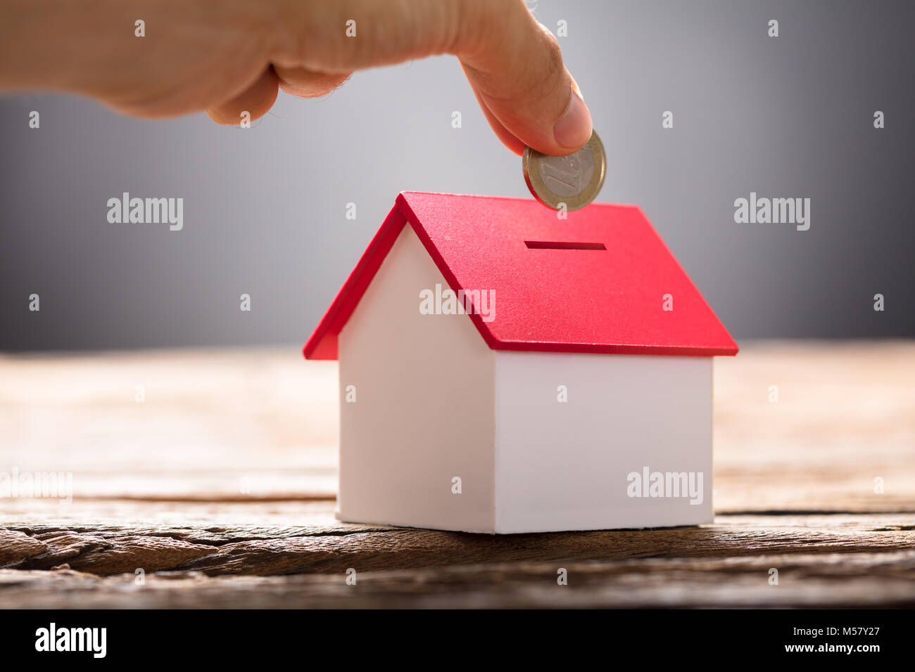 Closeup of hand putting coin in house piggy bank on wood Stock Photo