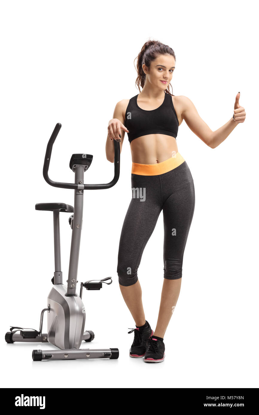 Full length portrait of a teenage girl leaning on a cross-trainer machine and making a thumb up gesture isolated on white background Stock Photo