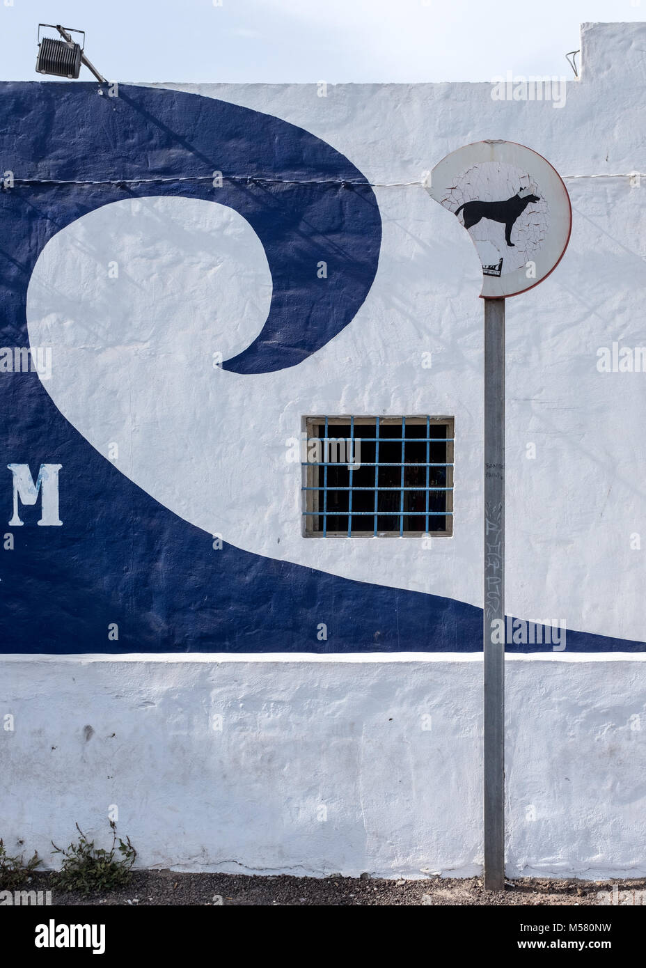 Bleached 'no dogs' sign in front of a wall with a barred window and a painted blue wave on it in Corralejo, Fuerteventura, Spain, Europe, Africa. Stock Photo