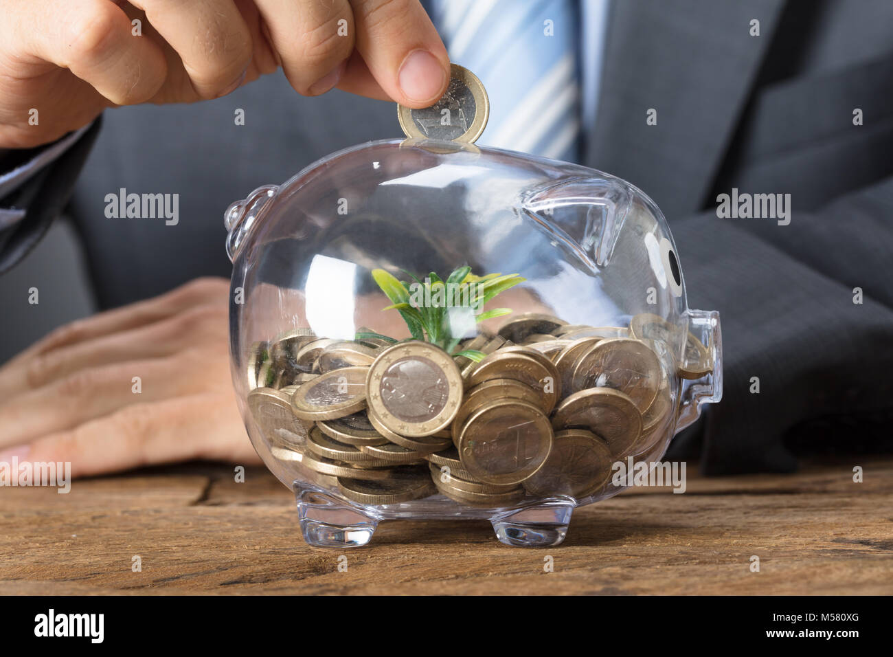 Closeup of businessman putting coin into transparent piggybank with plant Stock Photo
