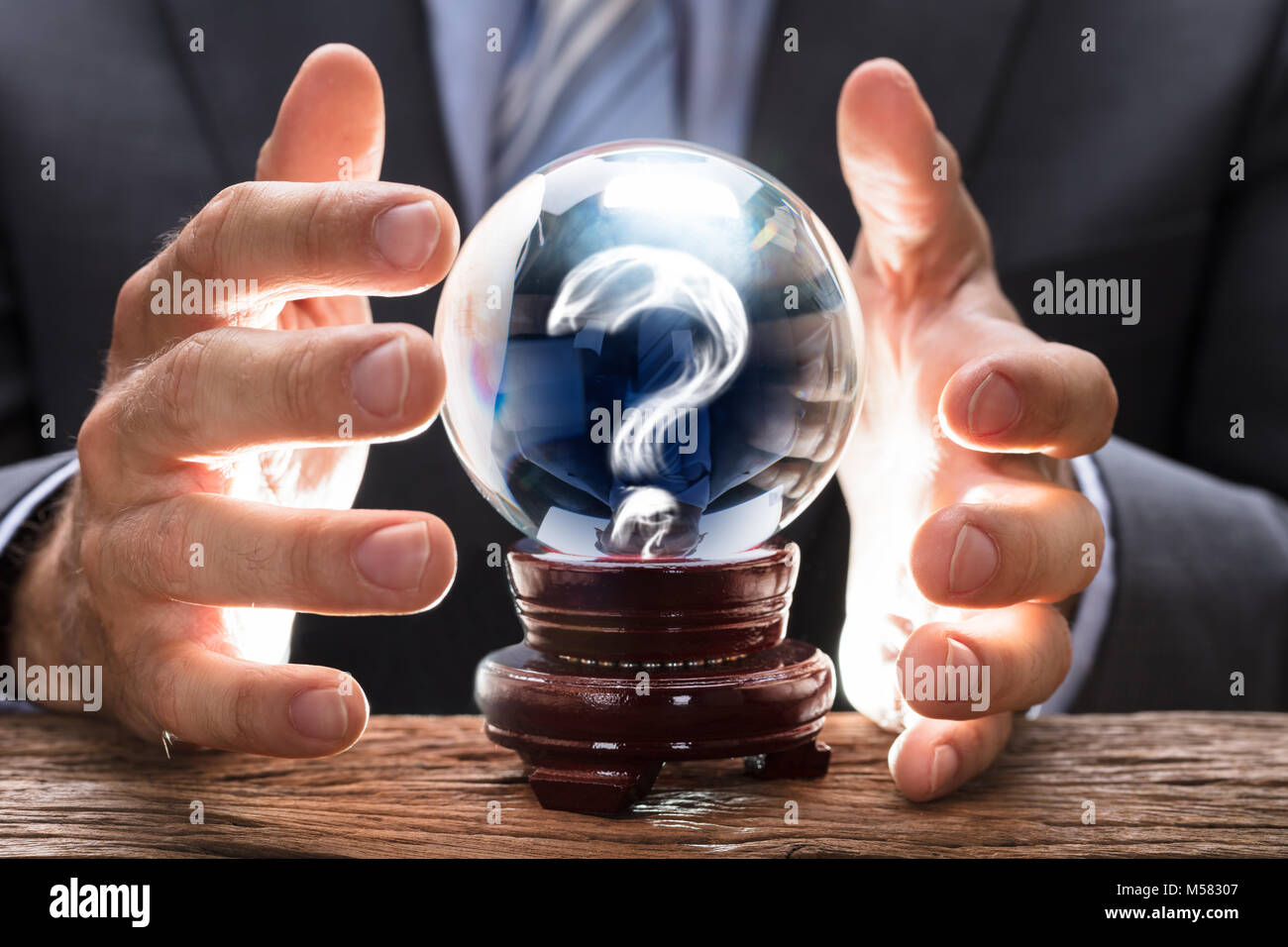 Midsection of businessman covering crystal ball with smoke forming question mark at wooden table Stock Photo