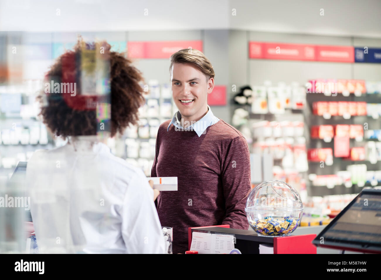 Portrait of a handsome young man smiling while buying an useful pharmaceutical product in a modern drugstore with various medicines and helpful person Stock Photo