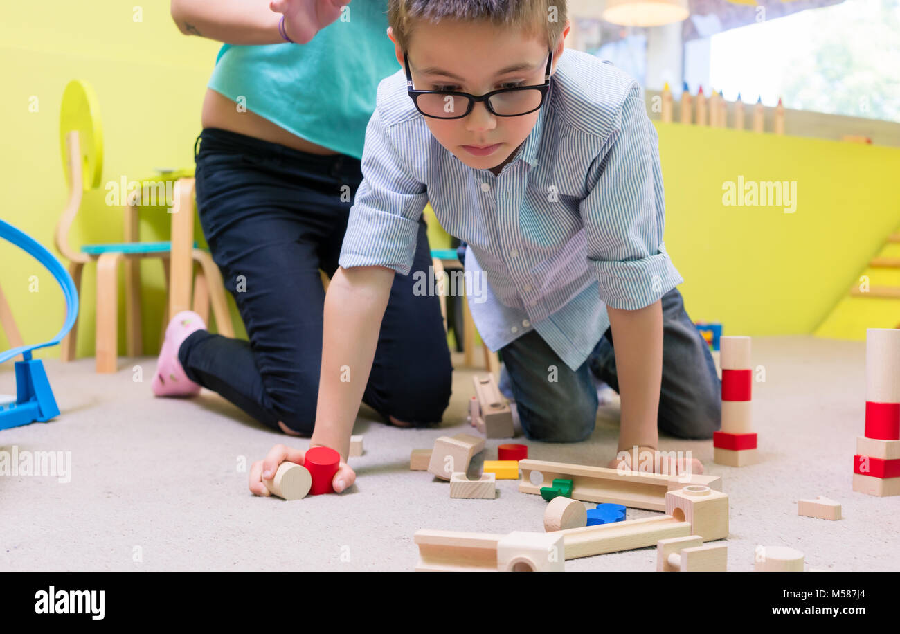Cute pre-school boy wearing eyeglasses while playing with wooden toys during supervised free playtime at the kindergarten Stock Photo