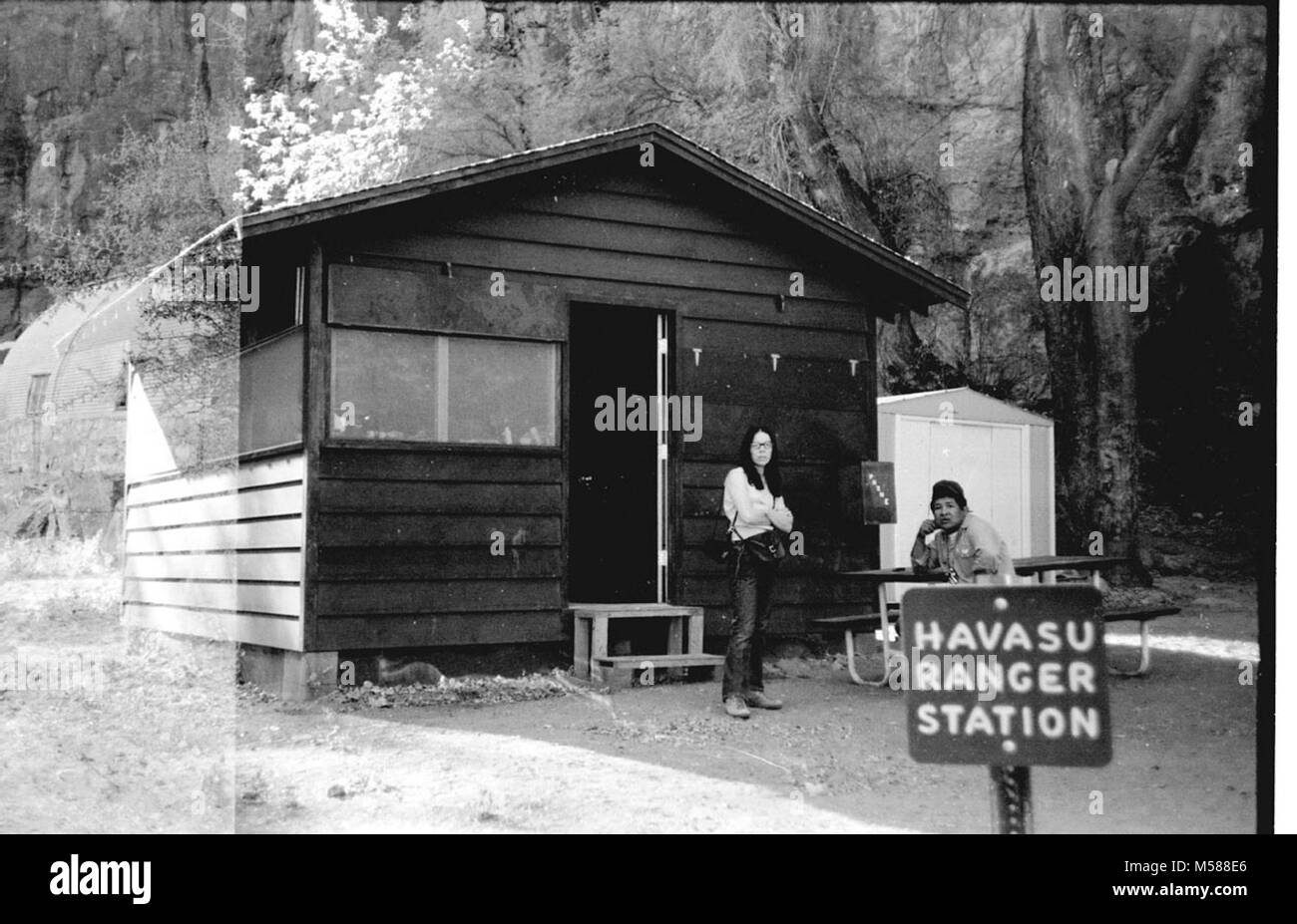 Grand Canyon Historic. EXTERIOR OF RANGER STATION. WOMAN STANDING. SUPAI EMPLOYEE OF NPS  CAMPGROUND AT HAVASU HAVASUPAI WHEN ADMINISTERED GRCA GRAND CANYON NATIONAL PARK. LEFT-SIDE DOUBLE EXPOSURE WITH SUPAI CHURCH ?  CIRCA 1977, Stock Photo