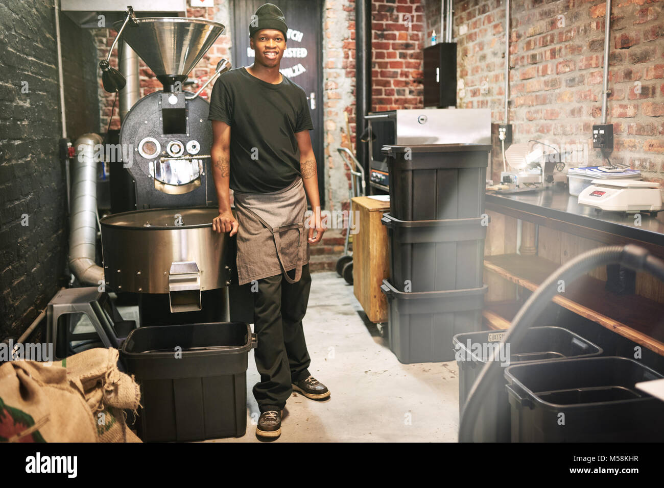 African worker standing in a coffee roasting room next to a coffee bean roaster in an industrial setting amongst all the other stored coffee beans and Stock Photo