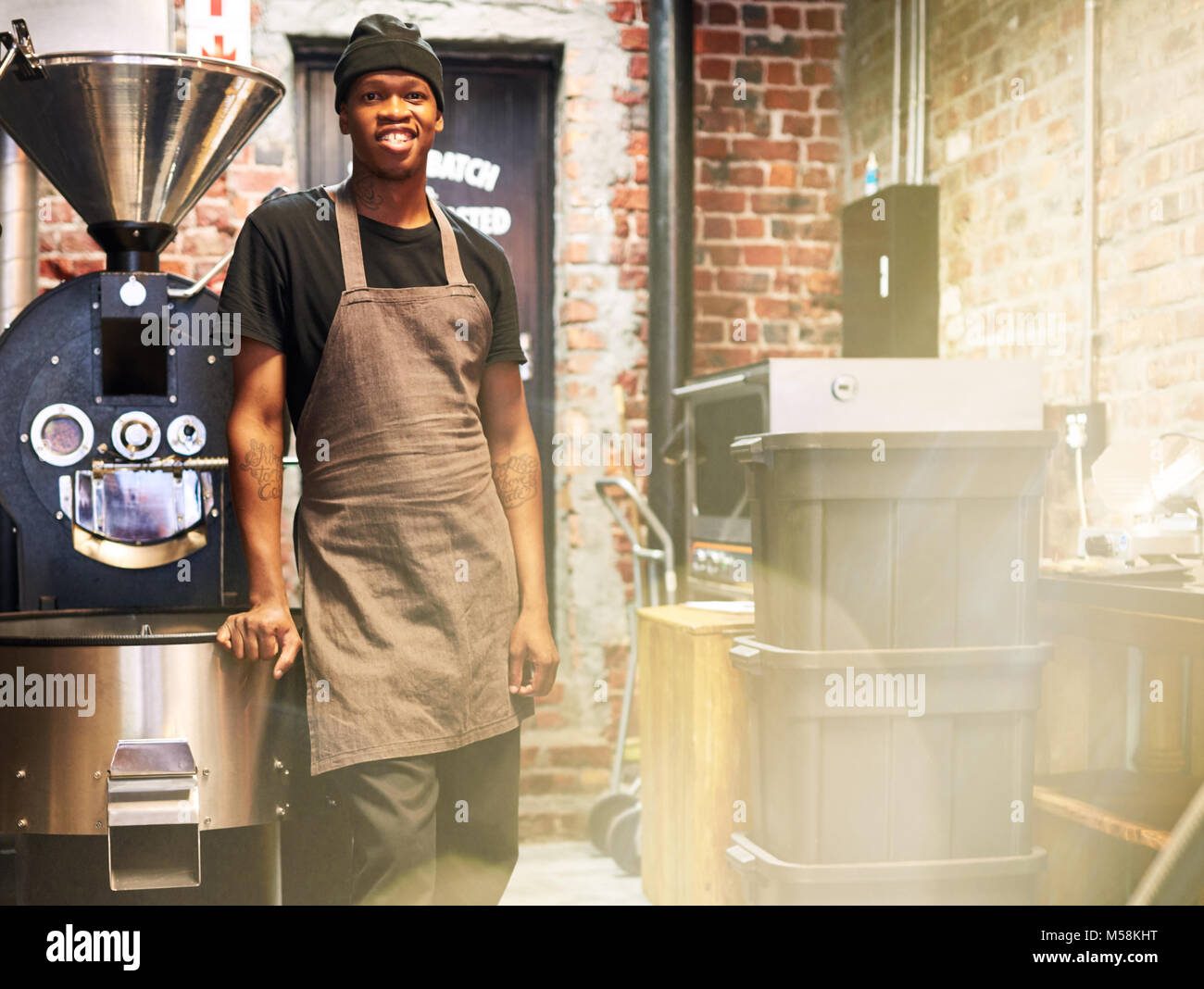African male employee smiling at camera while standing next to a coffee roasting machine amongst some other machinery to do with the making of coffee. Stock Photo