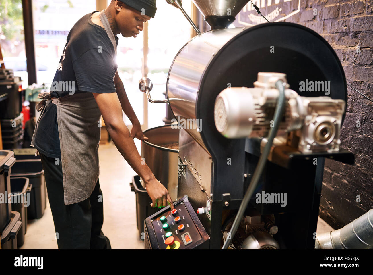 African man pushing red button on an industrial coffee machine, with large windows in the background allowing ample light into the space from behind t Stock Photo