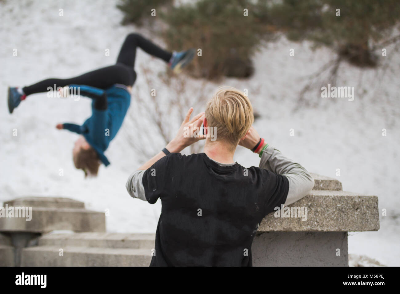 Blonde hair teenager makes photo on smartphone of acrobatic jump girl in winter city park - parkour concept Stock Photo