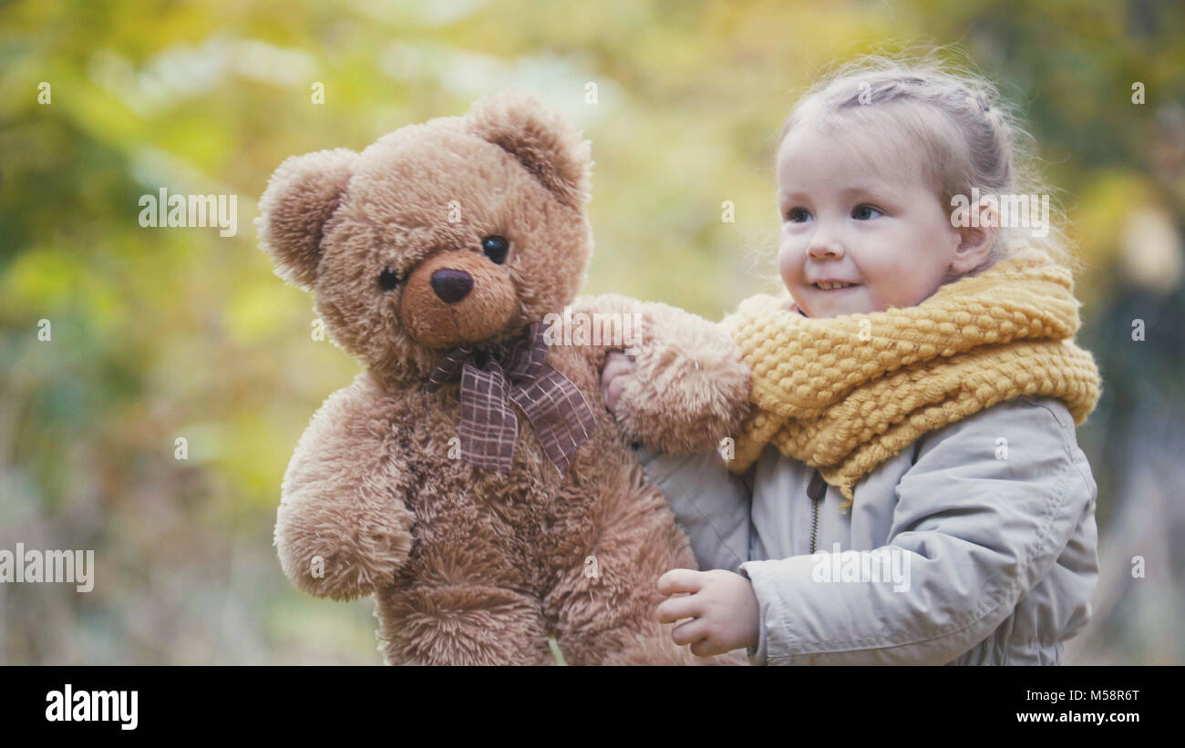 Little blonde girl plays with teddy bear in autumn park Stock Photo