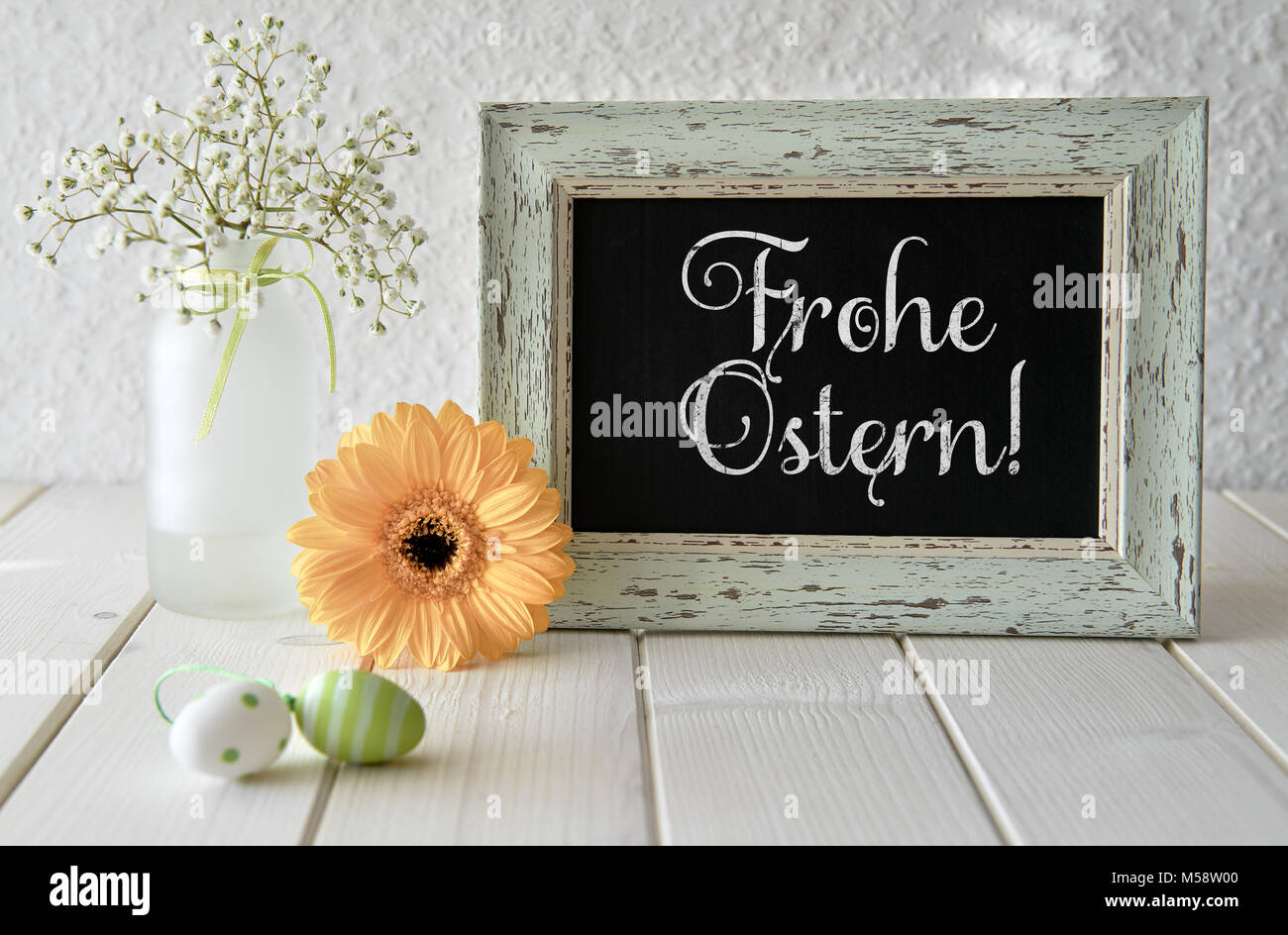 Spring flowers, Easter decorations and a blackboard on white table. Text 'Frohe Ostern', that means 'Happy Easter!' in English on a blackboard. Stock Photo