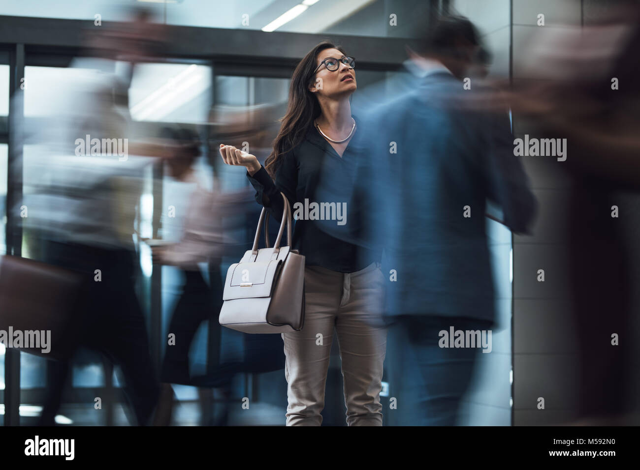 Businesswoman standing and looking upwards while waiting for someone with people rushing in the lobby. Motion blur effect. Stock Photo