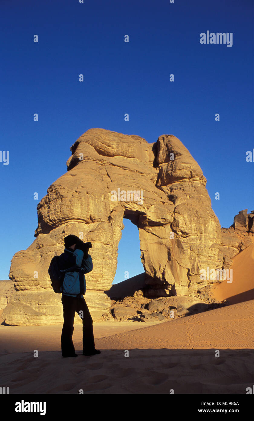 Libya. Near Ghat. Sahara desert. Akakus (Acacus) National Park. Natural arch called Fezzenger. Tourist, woman photographing arch. Stock Photo