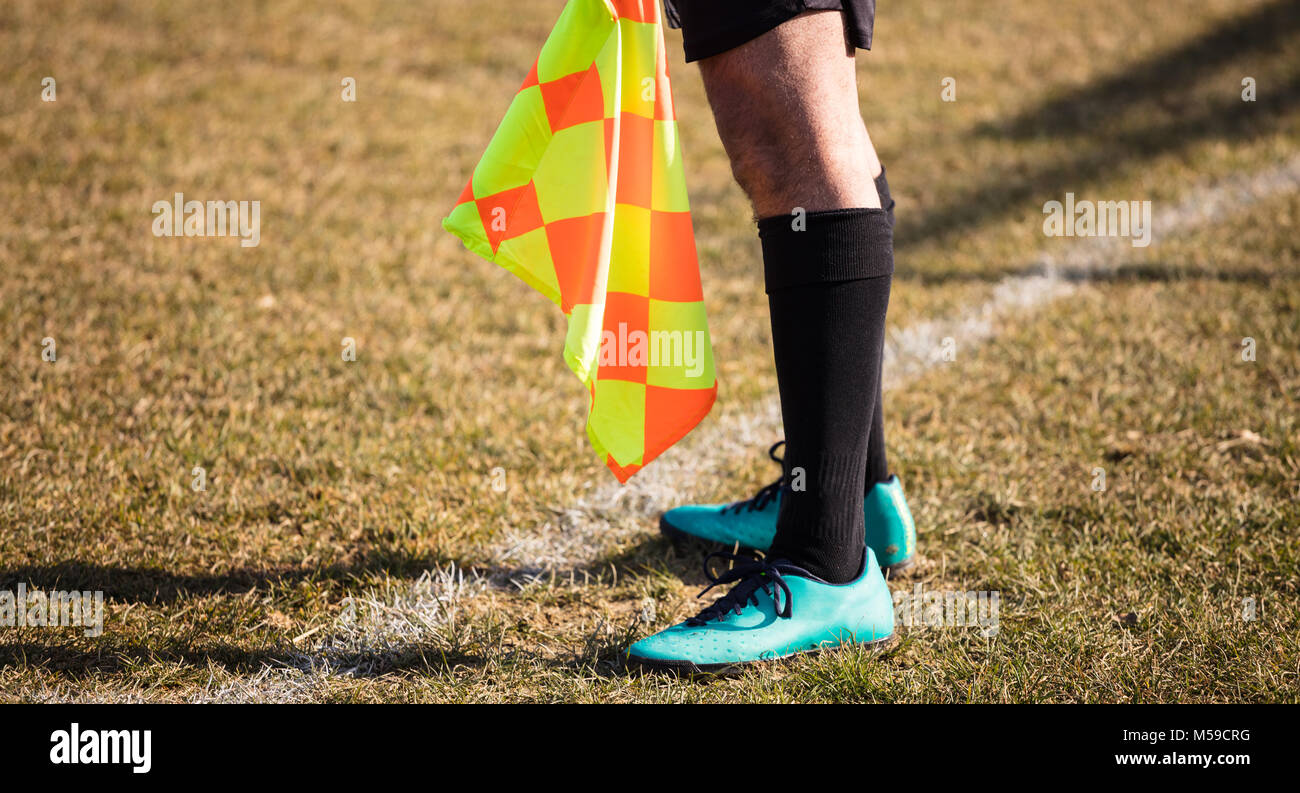 Football soccer arbiter assistant stands at sideline observing the match with flag at hands. Blurred green field background, close up. Stock Photo