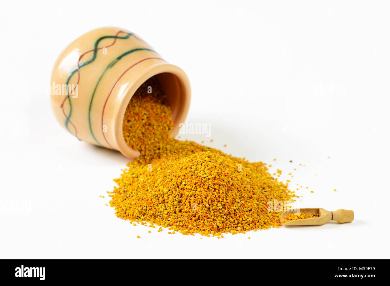 Heap of bee pollen granules, a wooden scoop and an overturned glazed clay pot full of pollen. Selective focus. Closeup. Stock Photo