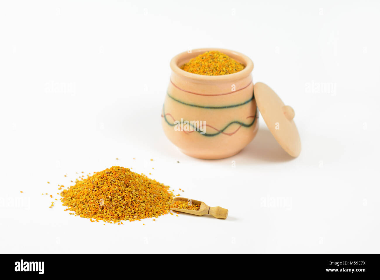 Pile of bee pollen and a little wooden scoop. In a blurry background, it is visible glazed clay pot full of pollen with a lid leaning against the pot. Stock Photo