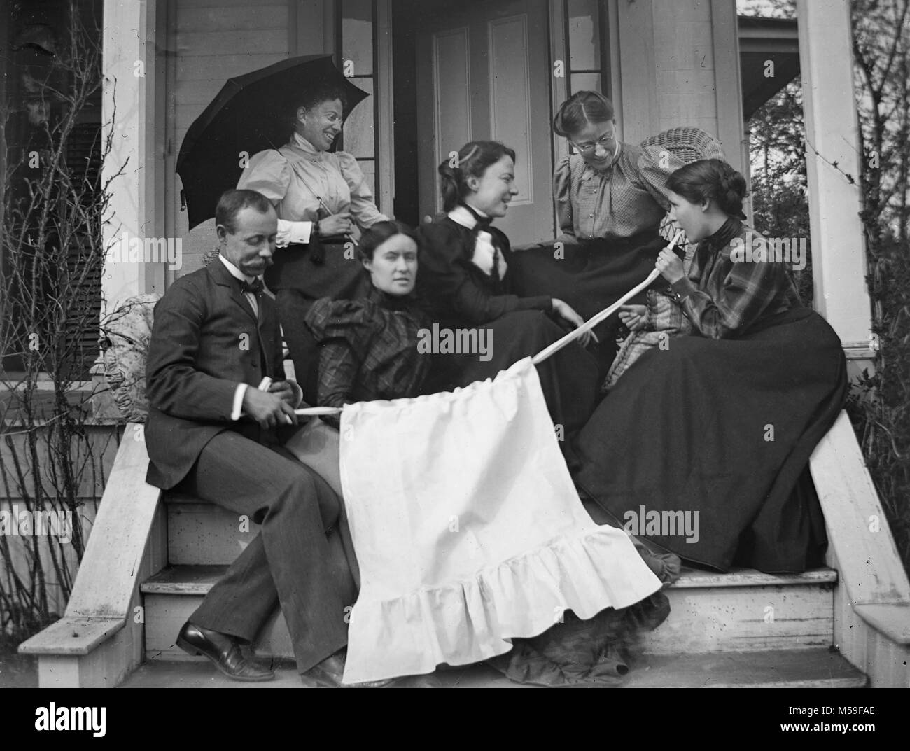 Wacky Victorians appear to hide a child behind an apron for a photo on the front porch, ca. 1900. Stock Photo