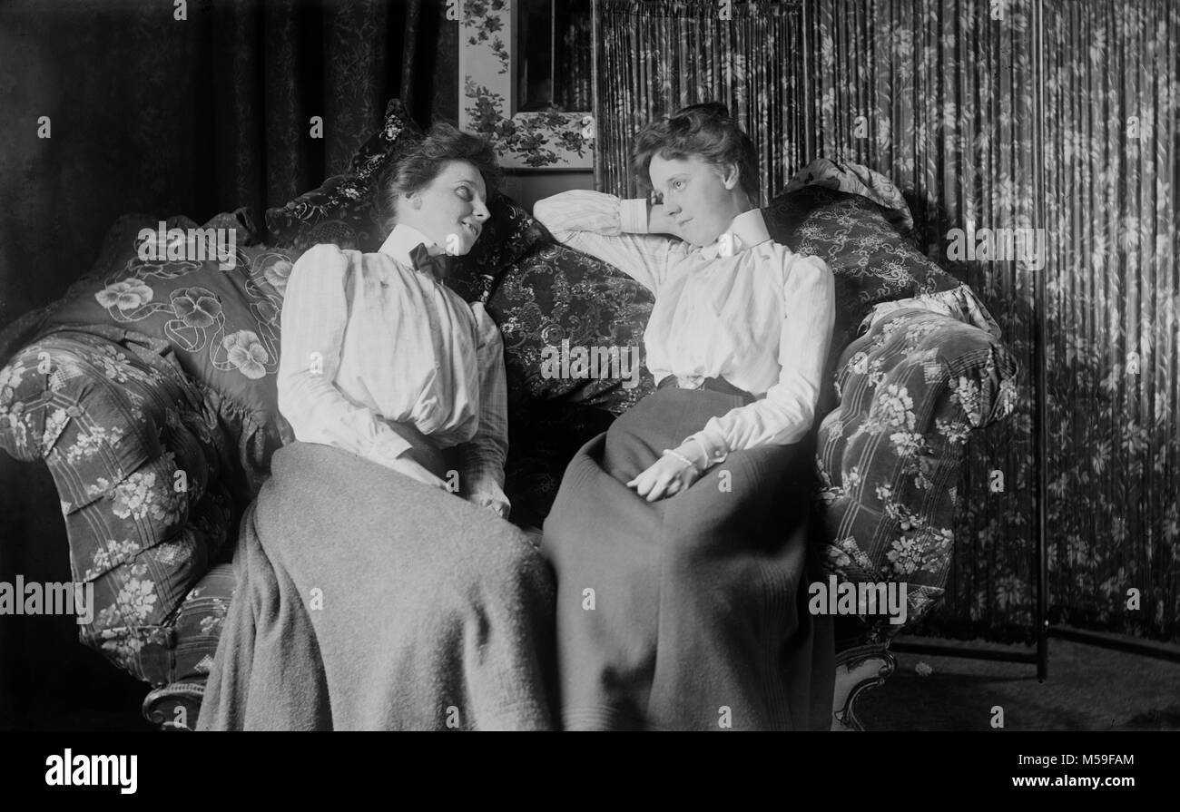 Two Victorian women lounge on an overstuffed couch together, ca. 1900. Stock Photo