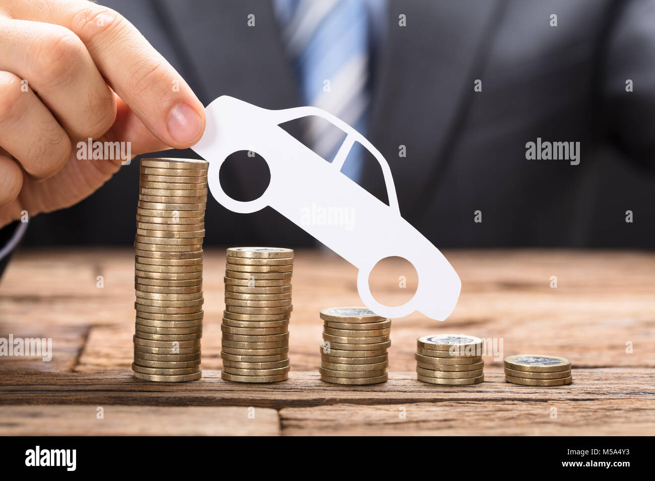 Businessman holding paper car on stacked coins arranged in declining order on wooden table Stock Photo
