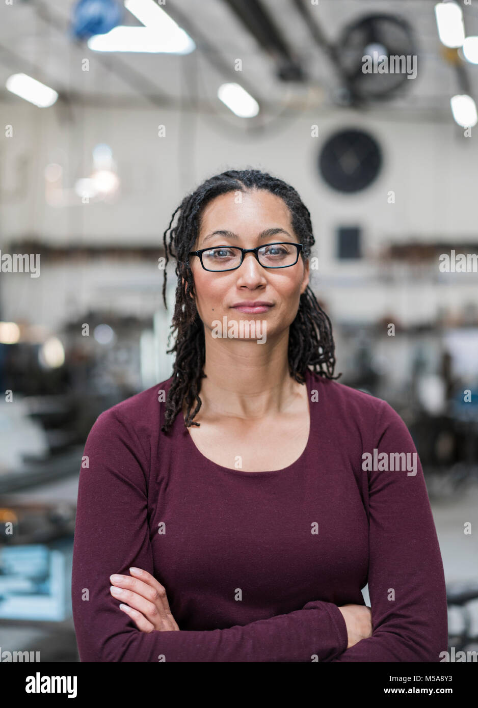 Woman with brown hair wearing glasses standing in metal workshop, smiling at camera. Stock Photo