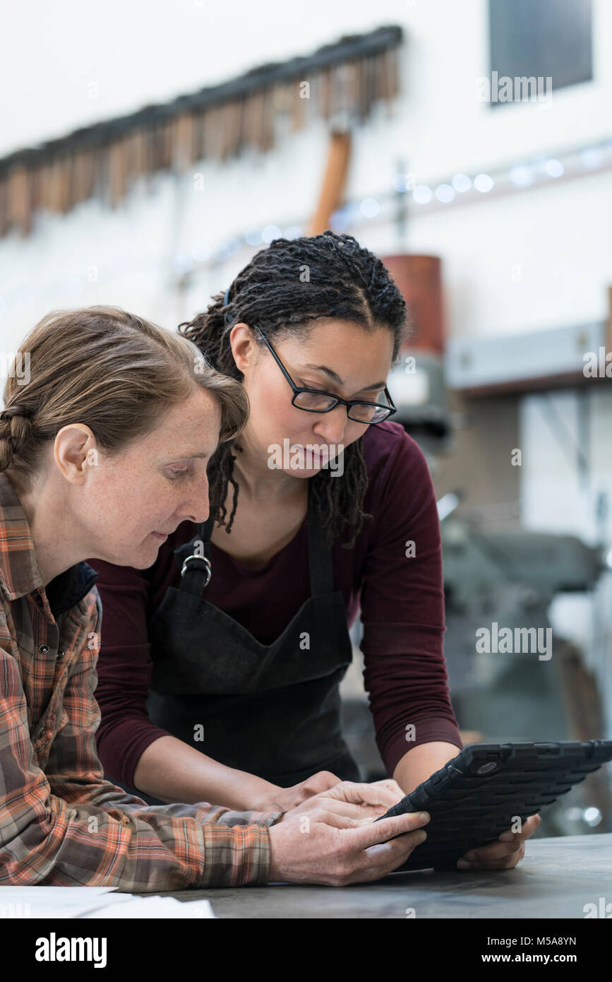 Two women standing at workbench in metal workshop, looking at digital tablet. Stock Photo
