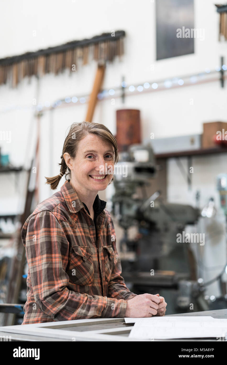 Blond woman wearing checkered shirt standing in metal workshop, smiling at camera. Stock Photo