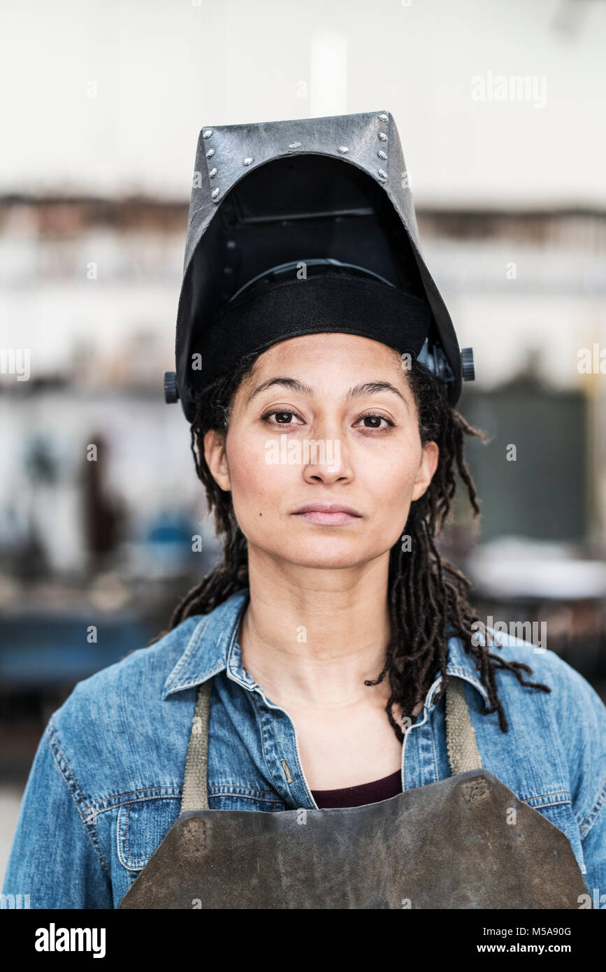 Portrait of woman wearing apron and welding mask standing in metal looking at camera