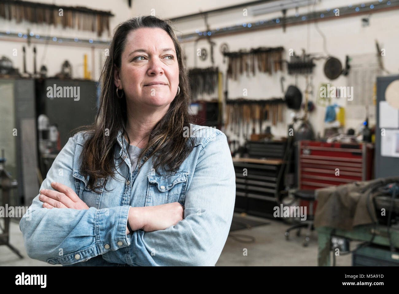 Woman with brown hair wearing Denim shirt standing in metal workshop, arms folded, smiling. Stock Photo