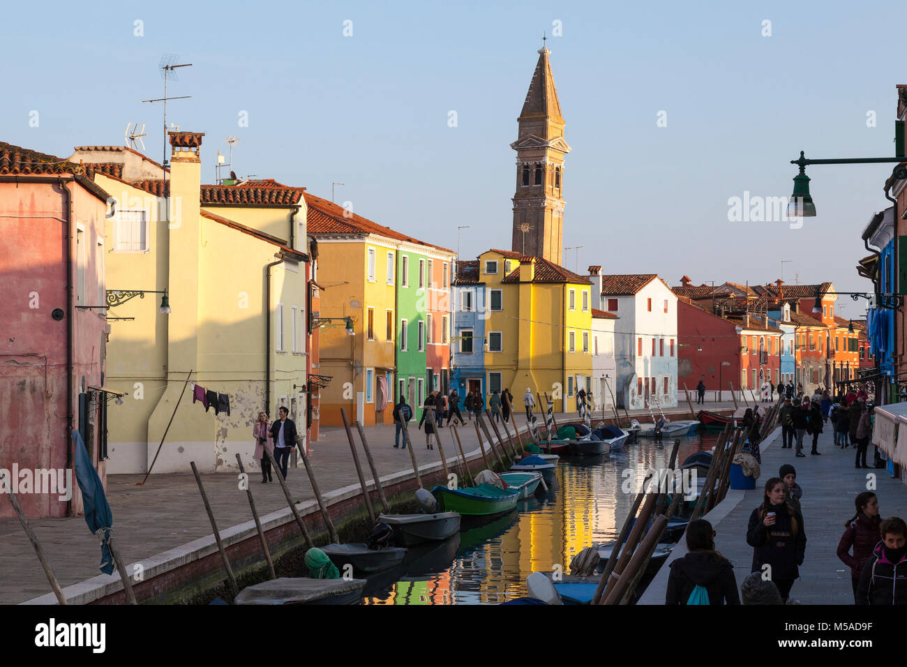 The bell tower of San martino Church at sunset, Burano Island, Venice,Veneto, Italy with reflections on the canal and people walking on Fondamenta Pes Stock Photo